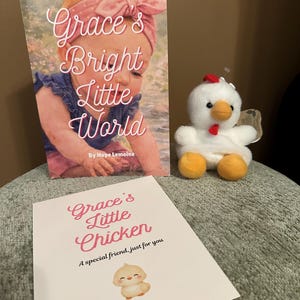 May include: A children's book titled "Grace's Bright Little World" with a watercolor illustration of a baby wearing a pink headband. A plush white chicken toy with orange feet and a red comb sits next to the book. A card reads "Grace's Little Chicken, A special friend, just for you."