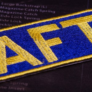 May include: A blue and gold embroidered patch with the letters "AFT" in gold.
