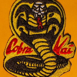 May include: Yellow and black embroidered patch with a cobra snake design and the text "Cobra Kai" in red.
