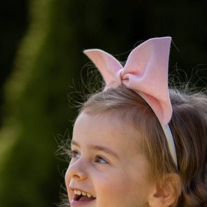 May include: A young girl with long, curly blonde hair is wearing a pink bow headband and a pink ruffled dress. She is smiling and looking up.