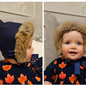 May include: A baby wearing a blue felt hat with brown fur ear flaps and a navy blue dress with orange floral print.