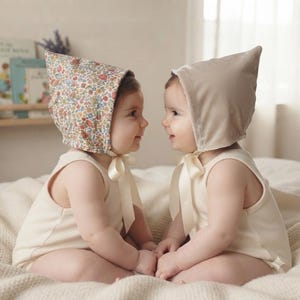 May include: Two infants wearing sleeveless cream-colored rompers and pointed bonnets. One bonnet features a colorful floral pattern, while the other is a solid light beige velvet. Both bonnets are secured with matching cream-colored ribbon ties.
