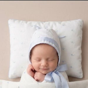 May include: A white baby bonnet featuring a light blue satin ribbon trim and tie, decorated with a subtle blue floral pattern. The bonnet is positioned on a matching white rectangular pillow with a light blue floral print, providing a soft accessory.