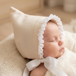 May include: A cream-colored bonnet with a pointed top, featuring a white ruffled trim along the face opening and a white ribbon tie under the chin. The bonnet is made of a soft, smooth fabric and is worn by a sleeping infant wrapped in a blanket.