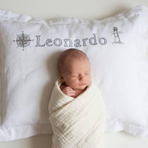 May include: A white rectangular pillowcase featuring embroidered dark gray line art of a compass rose on the left, the name Leonardo in the center, and a lighthouse on the right. The fabric has a visible texture and a stitched border edge.