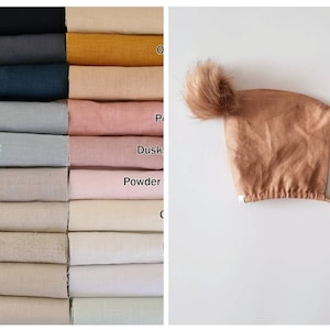 May include: A brown linen baby bonnet with a fluffy pom pom on top. The bonnet has a tie at the back. The bonnet is shown against a white background.