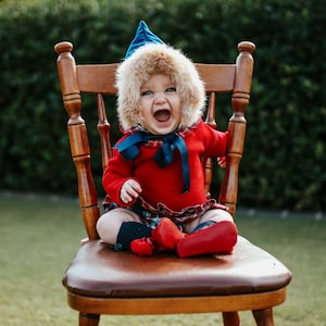 May include: A baby wearing a red long-sleeved jumper with a plaid collar, a matching plaid skirt, and a blue pointed hat with a tan faux fur trim. The outfit includes a dark blue ribbon tied under the chin and bright red shoes with pom-poms.