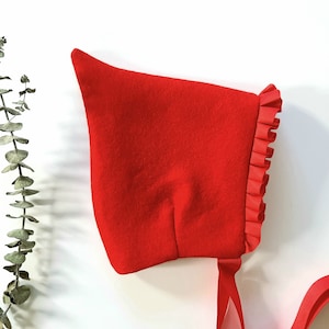 May include: A red felt bonnet with a ruffled edge and a red satin ribbon tie.