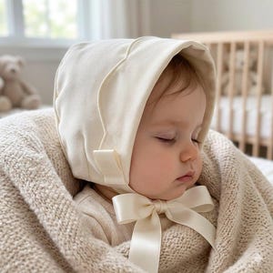 May include: A cream-colored bonnet made of a ribbed fabric featuring a structured brim and a smooth satin ribbon that ties into a bow under the chin. The bonnet is worn by a sleeping infant wrapped in a matching cream-colored knit blanket.