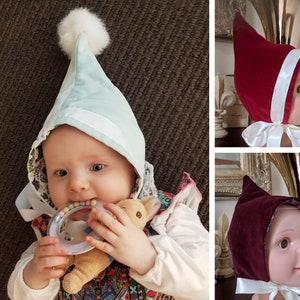 May include: Three baby bonnets in different colours: light blue, red and burgundy. Each bonnet has a white satin ribbon tied under the chin. The bonnets are made of soft fabric and have a pointed top.