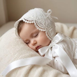 May include: A white infant bonnet featuring intricate floral lace embroidery along the scalloped edge and a smooth satin ribbon tie under the chin. The bonnet is crafted from a lightweight fabric and includes a decorative bow detail on the side.