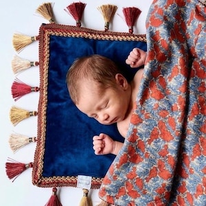 May include: A square blue velvet blanket with a decorative gold braided border and alternating red and beige tassels along the edges. The blanket is partially covered by a second fabric featuring a dense, textured pattern of orange and blue floral shapes.