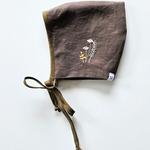 Puede incluir: Un gorro de bebé marrón con bordado floral. El gorro tiene un ribete y lazos verde oliva. El bordado presenta margaritas blancas y hojas verdes y doradas. Una pequeña etiqueta blanca con las letras "ld" está cosida en el lateral.