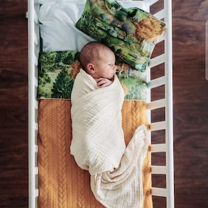 May include: A newborn infant wrapped in a cream-colored muslin swaddle blanket lies in a white crib. The bedding includes a mustard yellow cable-knit blanket and a pillow featuring a colorful forest landscape print with trees and deer.
