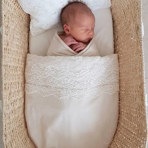 May include: A newborn infant wrapped in a cream-colored swaddle lies inside a woven wicker bassinet. The baby rests on a plain mattress with a white pillow featuring intricate lace trim, and a matching cream-colored blanket with lace detail.