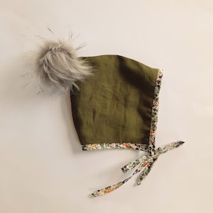 May include: A green baby bonnet with a white and floral trim and a grey fur pom pom.