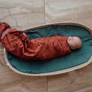 Puede incluir: Un bebé recién nacido está envuelto en un arrullo de muselina de color óxido con un pompón negro en la parte superior. El bebé está acostado en una cesta tejida forrada con una tela de color turquesa.