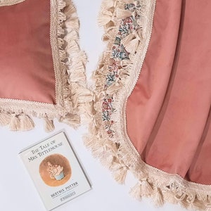 May include: Two pink velvet throw cushions with white fringe trim and tassels. The cushions are on a white surface with a book titled "The Tale of Mrs. Tittlemouse" by Beatrix Potter.