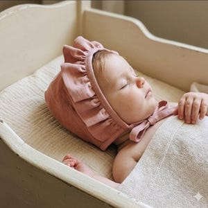 May include: A dusty rose bonnet featuring a ruffled trim along the front edge and a tie closure under the chin. The bonnet is made of a textured fabric and is worn by a sleeping infant who is partially covered by a light beige woven blanket.