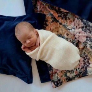 May include: A newborn infant with eyes closed is wrapped in a cream-colored knit swaddle blanket. The infant rests against a dark navy blue fabric and a patterned fabric featuring a floral design with shades of pink, orange, and dark green.