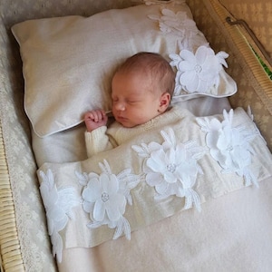 May include: A sleeping infant rests on a beige pillow adorned with white floral lace appliqués. The infant is covered by a matching beige blanket featuring a horizontal band of white floral lace appliqués across the center, set within a wicker bassinet.