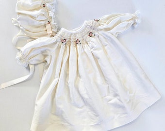 May include: A white smocked baby dress with a floral embroidery design and a matching white bonnet. The dress has puffed sleeves and a gathered skirt.