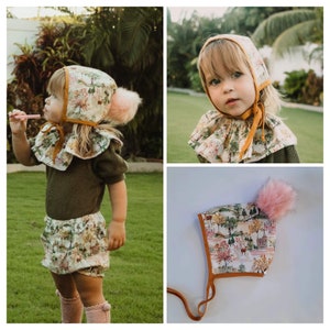 Puede incluir: Un gorro blanco con un pompón rosa y un ribete amarillo. El gorro tiene un estampado de escena de bosque. El gorro está atado con una cinta amarilla.