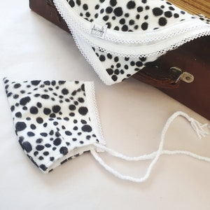 May include: A white and black dalmatian print fabric baby bonnet with white ties. The bonnet is lying on a white surface.