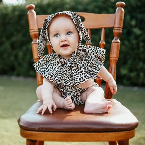 May include: A baby wearing a leopard print outfit consisting of a bonnet with long ear-like flaps and a matching ruffled collar and diaper cover. The fabric features a black, brown, and tan animal print pattern throughout the entire set.
