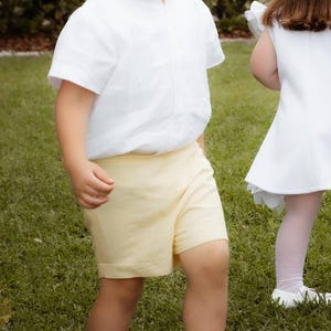 May include: A young child wearing a white short-sleeved shirt and yellow shorts. The child is also wearing white socks and white shoes. The child is standing on green grass. Another child is partially visible in the background.