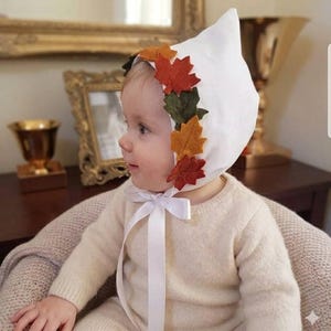 Puede incluir: Un gorro blanco puntiagudo con una hilera de hojas de arce de fieltro en tonos naranja, rojo y verde oscuro a lo largo del borde frontal. El gorro se sujeta bajo la barbilla con una cinta blanca, sobre un suéter de punto color crema de manga larga.