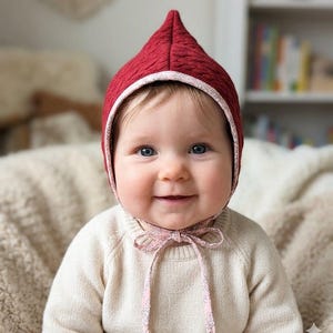 Puede incluir: Un gorro acolchado de color rojo intenso con la parte superior puntiaguda y un borde con estampado floral rosa claro. El gorro cuenta con lazos de tela floral rosa a juego que se atan bajo la barbilla, ofreciendo un ajuste seguro con textura.