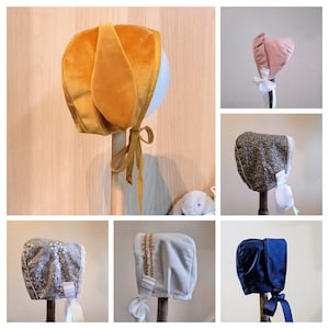 May include: A collection of six baby bonnets in various colours and fabrics, including velvet, satin, and floral print. The bonnets are displayed on mannequins with white ribbons.