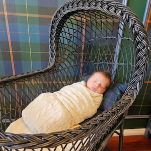 May include: A newborn infant is sleeping inside a dark brown wicker bassinet. The infant is wrapped in a cream-colored cable knit swaddle blanket. The bassinet features a high, arched hood with a woven pattern and a sturdy, open-frame structure.
