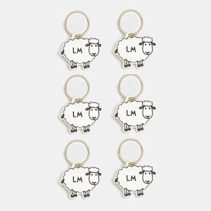 Pode incluir: Seis chaveiros com um desenho de ovelha branca, contornos pretos e as letras "LM". Cada chaveiro tem um anel prateado. As ovelhas têm uma aparência fofa e estão dispostas em um padrão de grade.