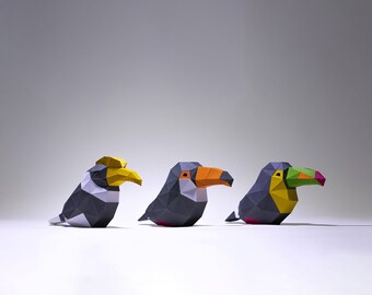 May include: Three origami birds, a toucan with an orange beak, a toucan with a green beak, and a hornbill with a yellow beak, are arranged in a row on a white surface.
