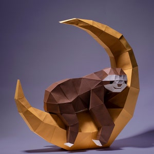 May include: A brown paper origami sloth is sleeping on a gold paper origami crescent moon. The sloth has a white face with black eyes and a small smile.