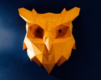 Pode incluir: Uma cabeça de coruja geométrica em papel, em tons de amarelo e laranja, montada numa parede azul escura. A face da coruja é composta por formas angulares de papel dobrado, criando um efeito tridimensional. Os olhos da coruja são escuros.