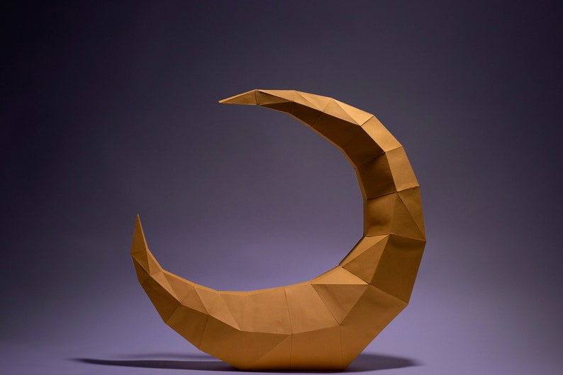 May include: A golden crescent moon sculpture made of paper, with a geometric, origami-style design. The sculpture is on a purple background.