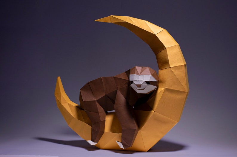 May include: A brown origami sloth is perched on a yellow origami crescent moon. The sloth has a white face and is positioned as if it is sleeping.