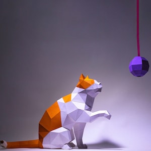May include: A white and orange paper cat figurine with a purple paper ball hanging from a string above it.