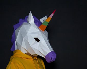 May include: A white paper mache unicorn mask with a purple mane and a rainbow horn. The mask is being worn by a person wearing a yellow hoodie.
