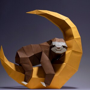 May include: A brown paper origami sloth is sleeping on a yellow paper origami crescent moon. The sloth has a white face with a black nose and mouth.