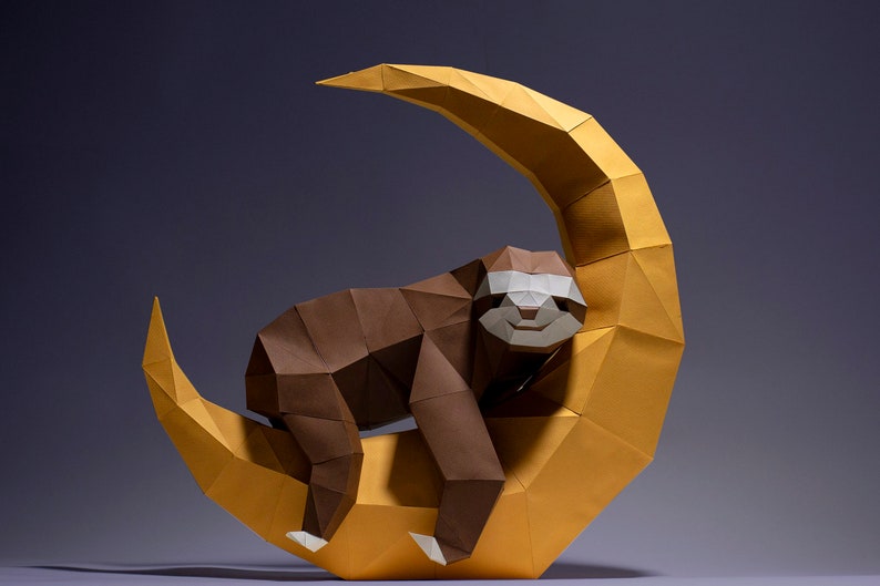May include: A brown paper origami sloth is sleeping on a yellow paper origami crescent moon. The sloth has a white face with a black nose and mouth.