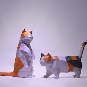 Op de afbeelding: Twee origami-katten gemaakt van papier. Eén kat is oranje, wit en zwart en staat rechtop. De andere kat is wit, oranje en zwart en ligt op de grond. Beide katten zijn gemaakt van gevouwen papier.