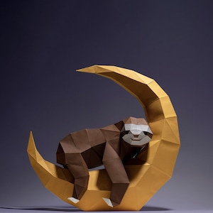 May include: A brown origami sloth is sleeping on a yellow origami crescent moon. The sloth has a white face with black eyes and a small smile.