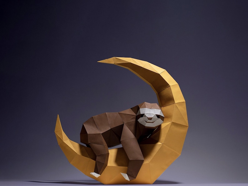 May include: A brown origami sloth is sleeping on a yellow origami crescent moon. The sloth has a white face with black eyes and a small smile.