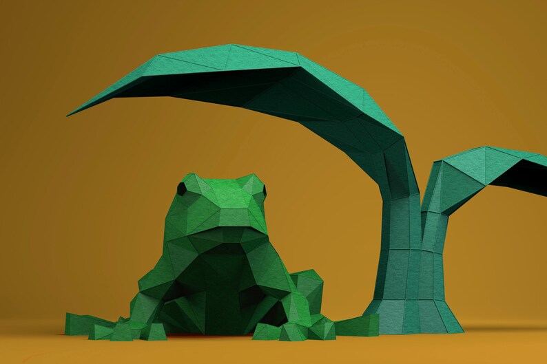 May include: A green origami frog sits in front of a green origami tree with two leaves. The frog and tree are on a yellow background.