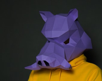Pode incluir: Uma máscara geométrica de papel roxo em forma de cabeça de javali. A máscara é usada sobre um moletom com capuz amarelo. A máscara tem facetas triangulares e está sobre um fundo cinza escuro.