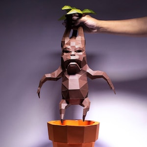 Pode incluir: Uma figura de papel castanho de uma criatura com um corpo humanoide e uma cabeça frondosa, de pé num vaso laranja. A figura é feita de papel dobrado e tem uma superfície detalhada e texturizada.